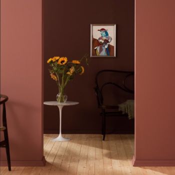 Valge ümmargune laud vaseõitega tumeroosal taustal, pruunis toonis seinad ning puidust tool ja põrand. Seinal on kunstiteos.