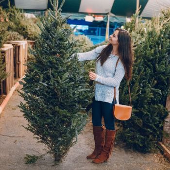 Naine valib jõulupuud, kandes pruune saapaid, siniseid teksaseid ja halli kampsunit, käes oranž kott.