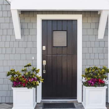 Musta värvi välisuks kahe lillevaasiga. Ukse kõrval on numbrimärk ja uksekell, asetseb hallide seinte vahel.