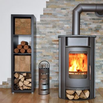 Hall metallist puuküttega ahi, leekidega, kõrval puiduhoidla ja väike metallist kaminapuude alus. Taustal kivi sein.
