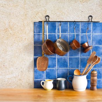 Sinine keraamiliste plaatidega seinapaneel köögis, millel on mitmesuguseid köögitarvikuid nagu potid, pannid ja lusikad riputatud ning väike valge kann ja mõned puidust maitseainepurgid laual.