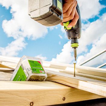 Akutrelliga puidule kruvi keeratav käsi, pilves taeva taustal. Nähtavad on puitdetailid ja kruvipakk.