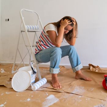 Naine istub remonditöökohas maha kukkunud valge värvipurgi kõrval, käed peas ja näos mure ilme. Ümbruses on segamini ehitusvahendid.