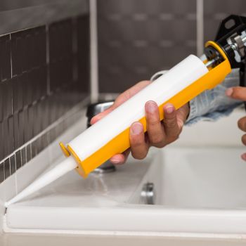 Käsi kasutab kollast tihenduspüstolit, et valget hermeetikut vannitoa valamukapi servale kanda.