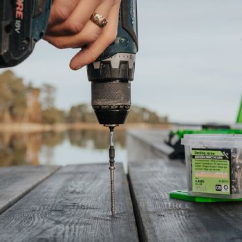 Käsi kasutab akutrelli puidupinna kruvimiseks, näha on ka kruvipakendit. Taust hägus.