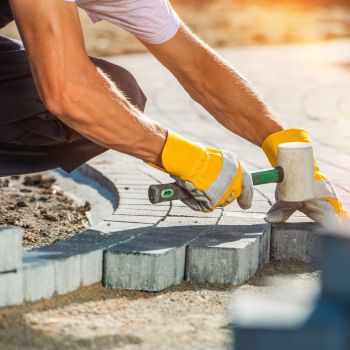 Töömees paigaldab halli tänavakivi, kasutades kollaseid kindaid ja elektrilist nurklihvijat.