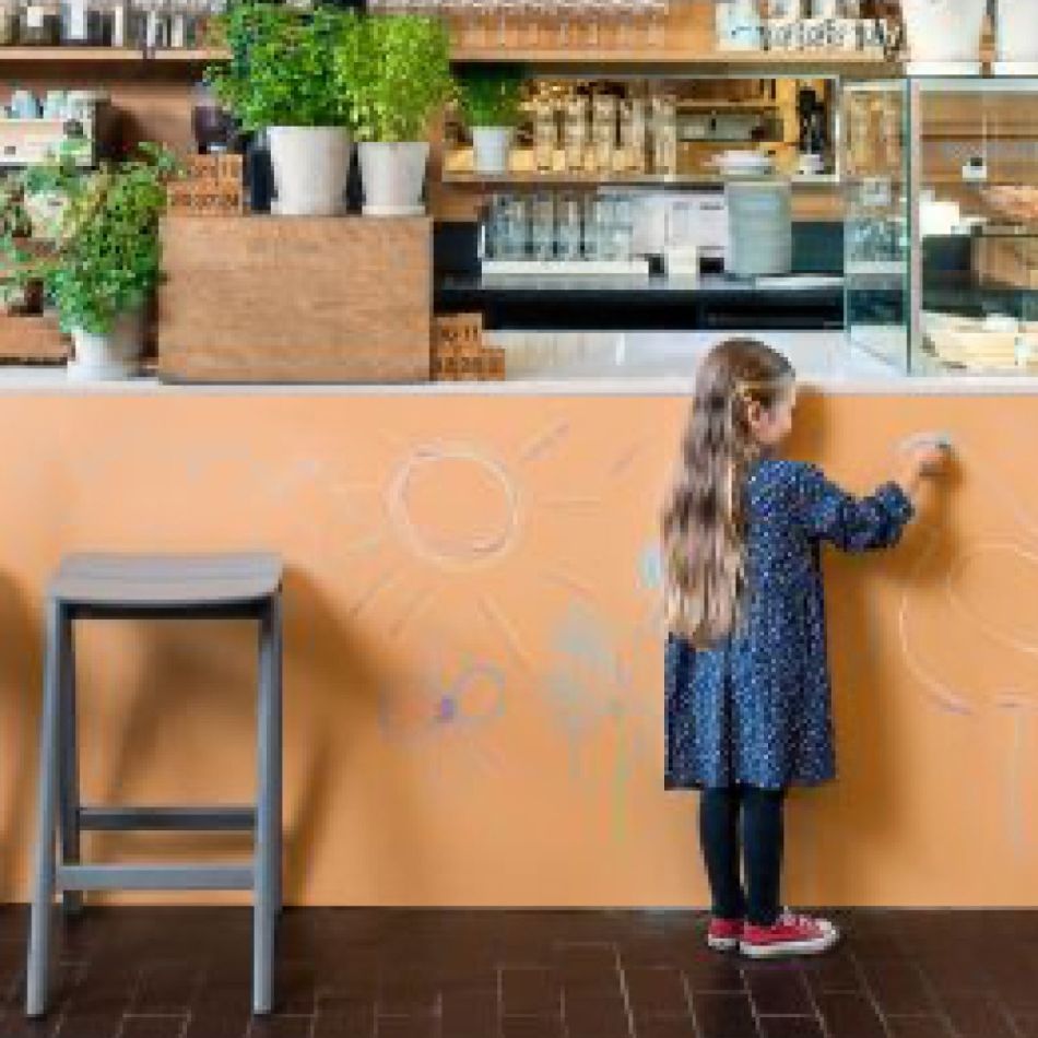 Tüdruk seisab kohvikus ja kirjutab kriidiga apelsinikarva seina peale. Taustal on puidust riiulid ja baarilett.