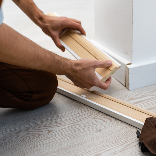 Mees paigaldab põrandaliiste, kasutades vasarat. Näha on puitpõrand ja valge seinaosa.