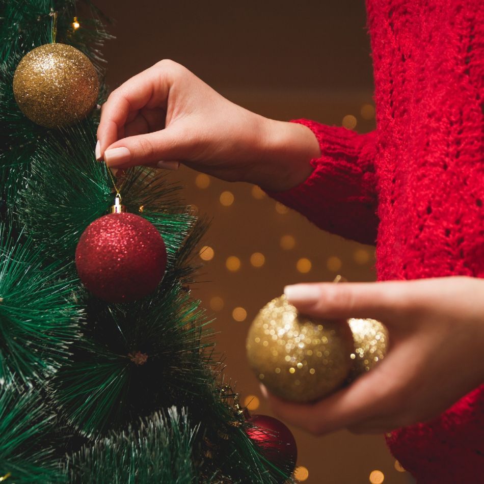 Isik ehib rohelist jõulupuud punaste ja kuldsete kuulidega, punases kampsunis käed.