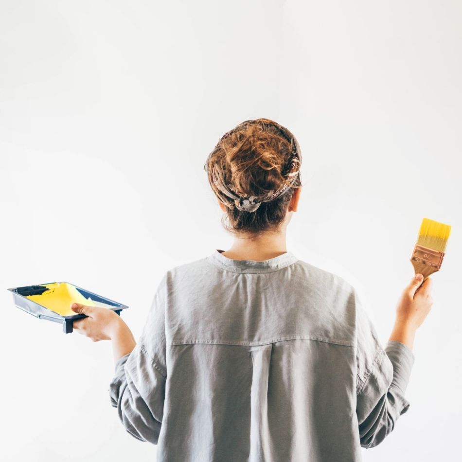 Naine hallis pluusis, hoiab käes kahte erinevat värvivalikut. Valget värvi ja kollast paletti kasutatakse illustreerimaks keskkonnasõbralike värvide valikut.