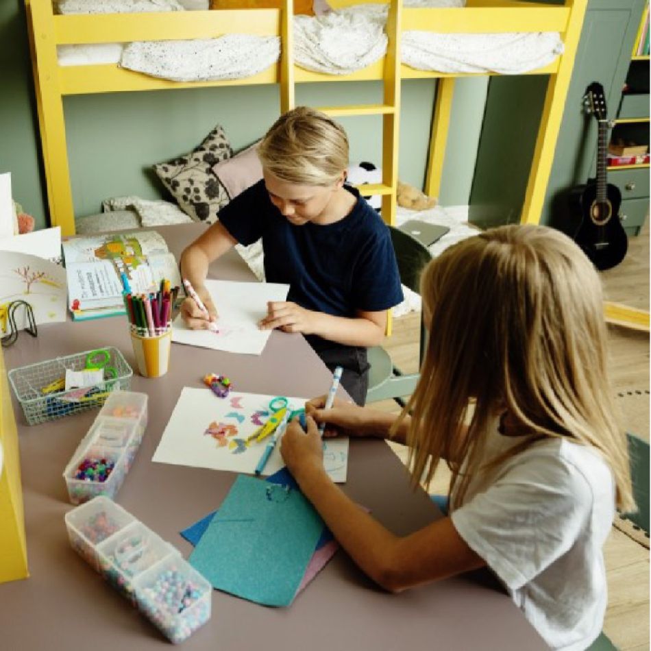 Kahe lapsega koolilapse tuba, kus on kollane kahe korrusega töölaud ja koolitarbed. Lapsed joonistavad ja kirjutavad.