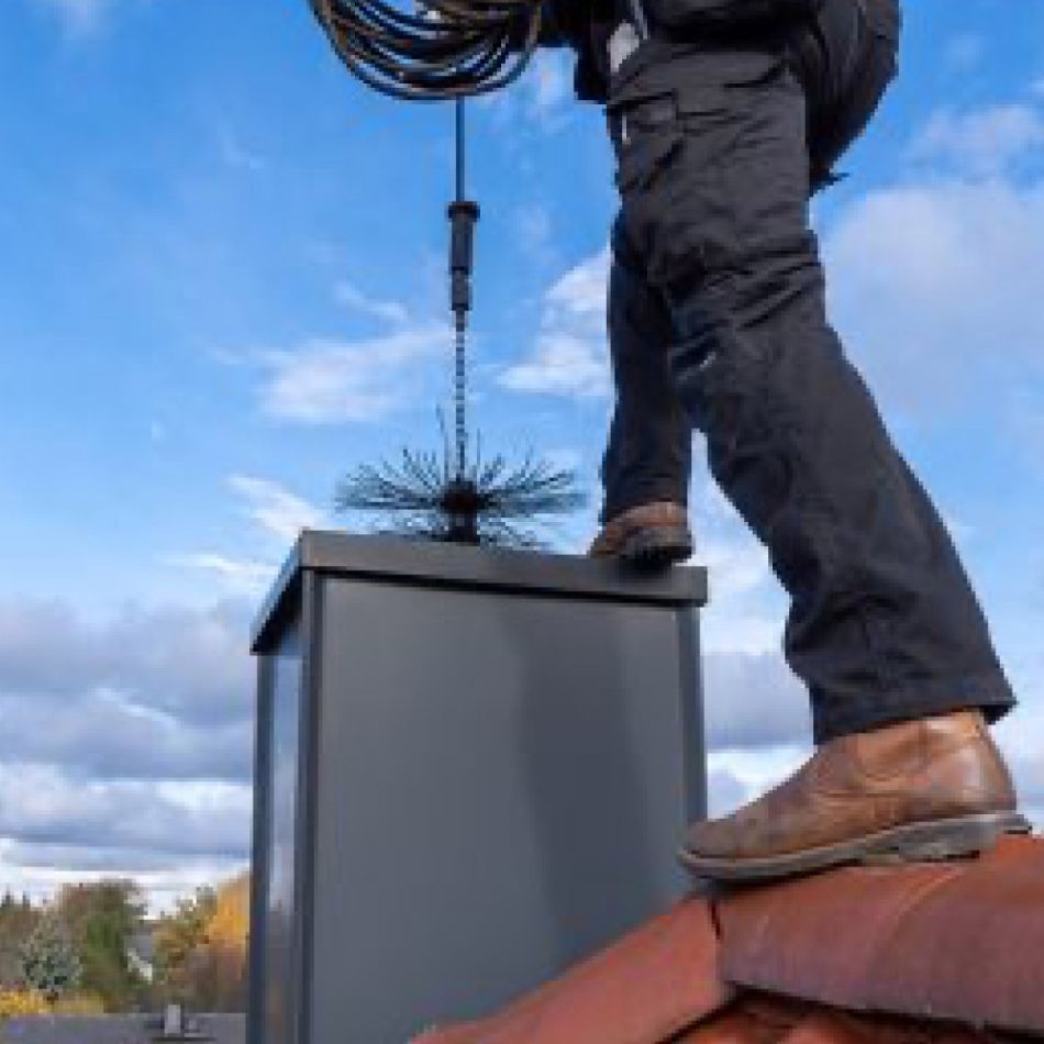 Mees puhastab korstent, kandes sinist tööriietust ja pruune saapaid. Ta seisab punasel katusekivil, taustal on pilvine taevas.