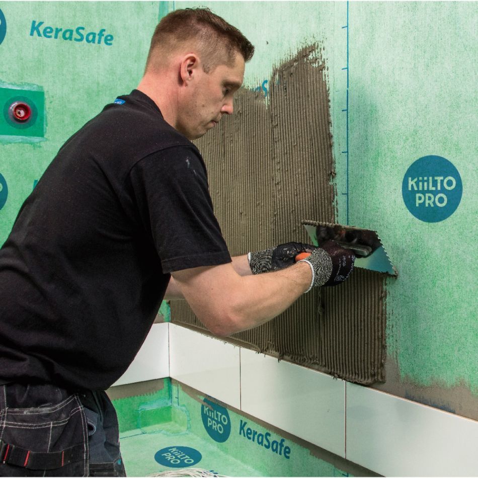 Mehe kujutis, kes kasutab hüdroisolatsioonimörti seina peale kandmiseks. Ta kannab halli T-särki ja mustrilisi pükse, tagataustal on roheline seinaisolatsiooni materjal näidistega.