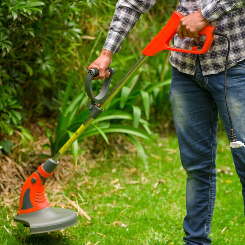 Aias trimmerdav mees, kasutades punast elektrilist rohutrimmerit. Trimmer on varustatud reguleeritava käepidemega. Taustal roheline taimestik.
