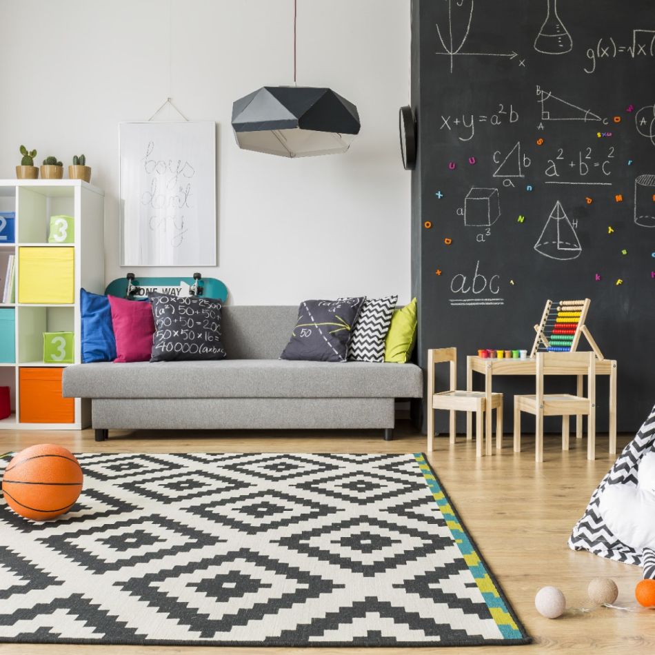 Kaasaegne lastetuba geomeetrilise mustriga vaiba, värvikate patjadega hallil diivanil ja musta tahvlivärviga sein, millel on joonistatud geomeetrilised kujundid ja matemaatilised valemid.
