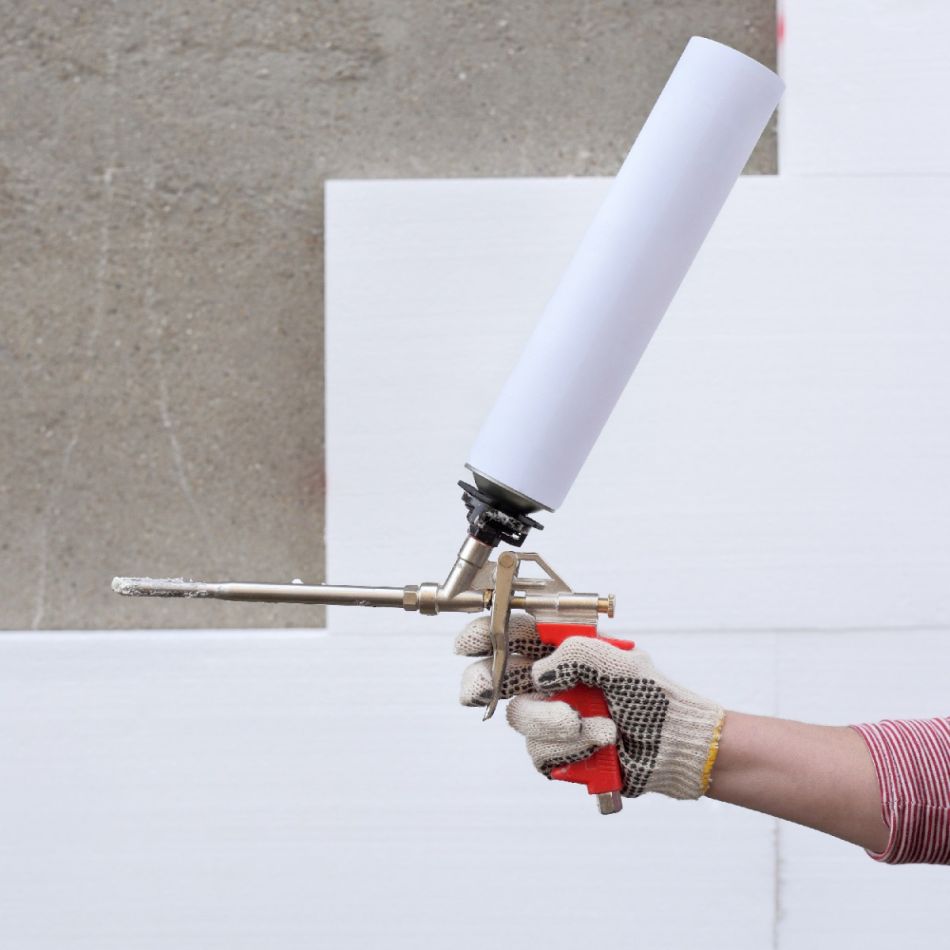 Käsi, mis hoiab liimipüstolit, süstides valget vahtu seina läbivasse torusse.