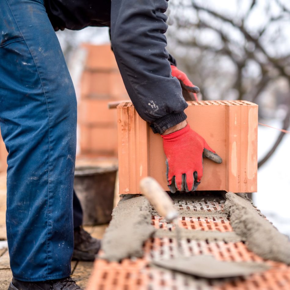 Person ehitab punastest tellistest seina, kandes siniseid tööpükse ja punaseid kummikindaid. Talveolud.