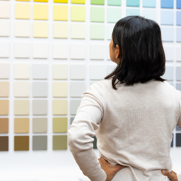 Naine seisab seljaga, vaadates erinevaid värviproove suurel seinal. Tal on seljas beež kudum ja ta hoiab käsi puusadel.