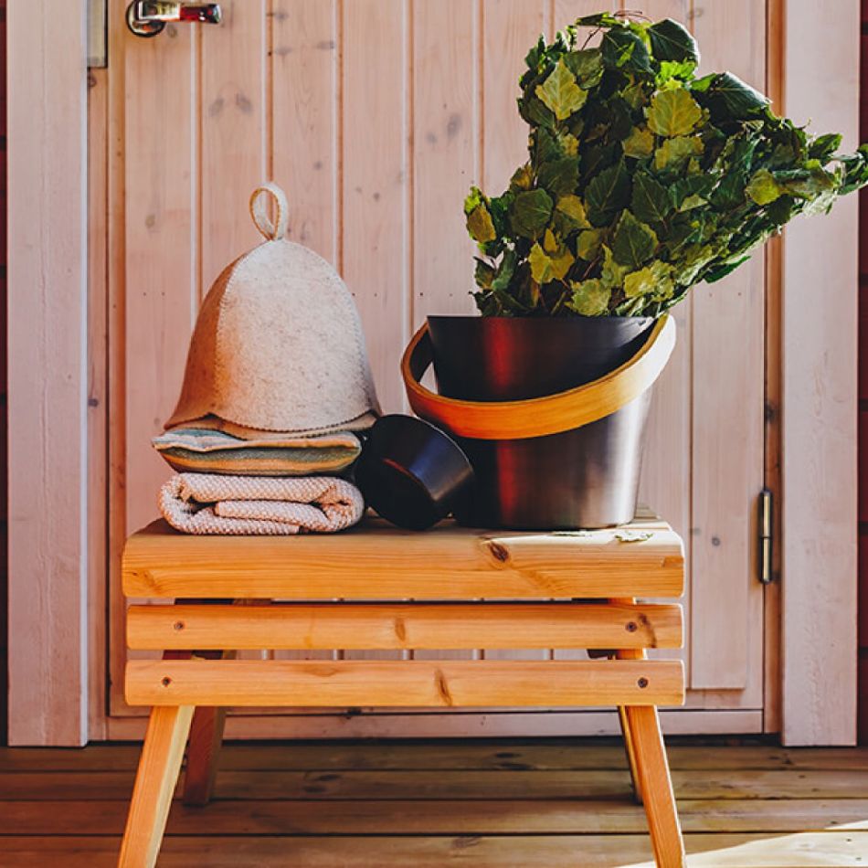 Puidust saunapink, millel on rätikud, saunamüts ja musta ning kollase ämbriga suur roheline taim.
