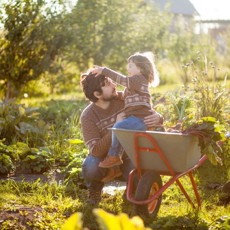 Pilt isast ja lapsest aias, laps istub aiakärus. Mõlemad naeratavad ja ilm on päikseline. Taustal roheline aed.