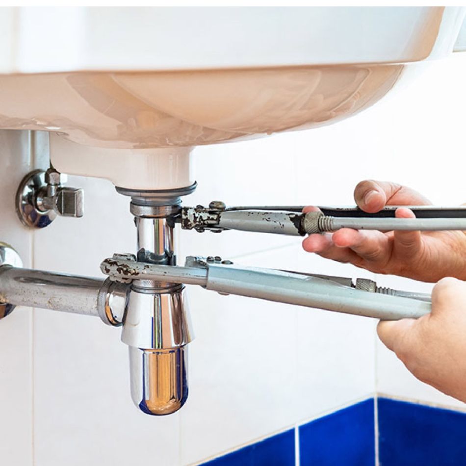 Käed paigaldavad valamu all torustikku, kasutades reguleeritavaid mutrivõtmeid ja kroomitud torusid. Taustal on siniselt plaaditud sein.